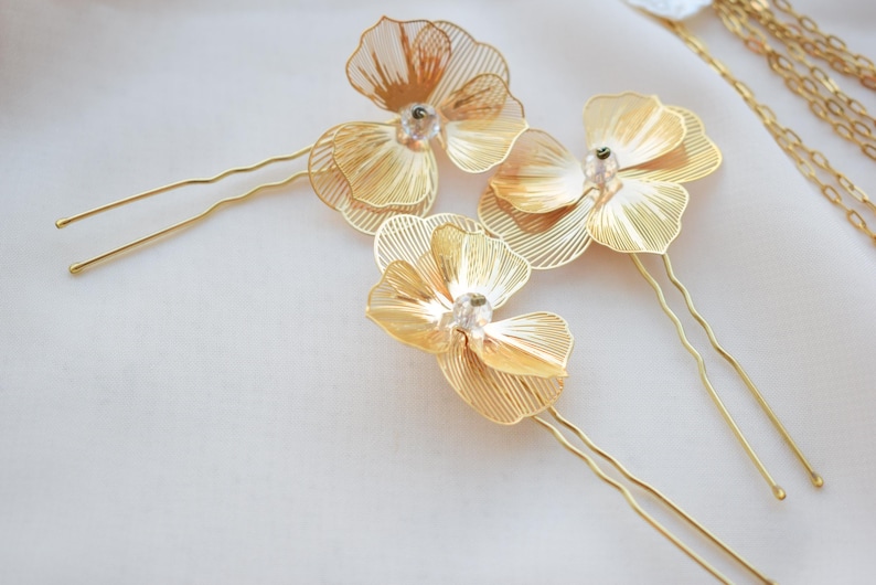 May include: Gold-toned metal hair pins with delicate, openwork flower designs. Each flower features a central crystal accent. The pins are arranged on a soft, white surface, suggesting a bridal or special occasion theme.