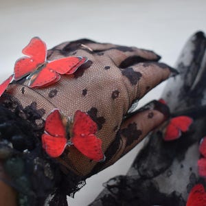 May include: Black lace gloves adorned with vibrant red butterfly appliqu&eacute;s. The gloves feature a delicate floral pattern and are designed to cover the hands. The butterflies add a whimsical touch to the elegant design.