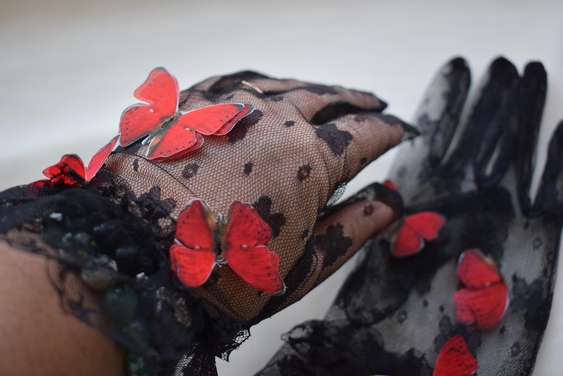 May include: Black lace gloves adorned with vibrant red butterfly appliqu&eacute;s. The gloves feature a delicate floral pattern and are designed to cover the hands. The butterflies add a whimsical touch to the elegant design.