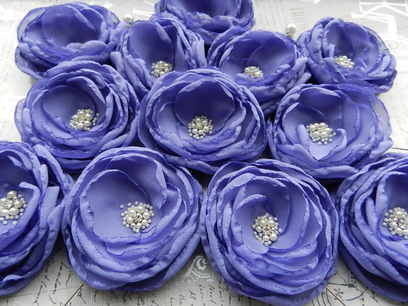 May include: A collection of 10 purple fabric flowers with pearl embellishments. The flowers are arranged in a circular pattern and have a soft, ruffled texture.