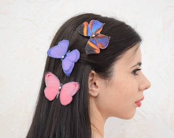 Puede incluir: Tres pinzas para el pelo con forma de mariposa en rosa, morado y azul con detalles en negro y naranja. Las pinzas se llevan en el pelo largo y oscuro de una mujer.