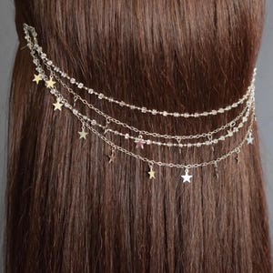Könnte beinhalten: Eine silberne Haarkette mit mehreren Strängen und sternförmigen Anhängern. Die Kette besteht aus kleinen Perlen und ist zum Tragen im Haar gedacht.
