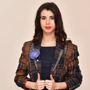 May include: A woman wearing a plaid blazer with a blue flower pin. The blazer has a brown, black, and white plaid pattern.