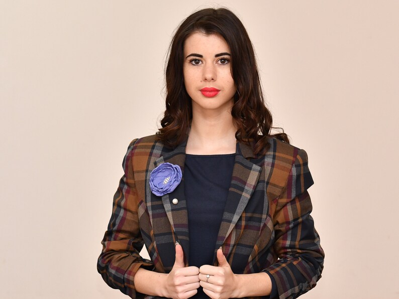 May include: A woman wearing a plaid blazer with a blue flower pin. The blazer has a brown, black, and white plaid pattern.