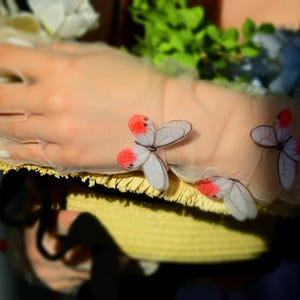 May include: A sheer, light pink glove adorned with delicate butterfly appliqu&eacute;s featuring white wings and red accents. The glove rests on a yellow straw hat, with floral accents in the background.