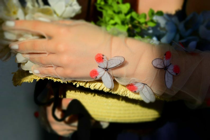 May include: A sheer, light pink glove adorned with delicate butterfly appliqu&eacute;s featuring white wings and red accents. The glove rests on a yellow straw hat, with floral accents in the background.