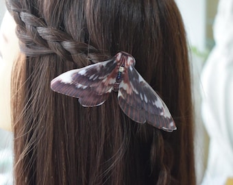 May include: A decorative hair clip shaped like a moth, featuring wings with shades of brown, white, and black. The clip is adorned with small, colorful beads and is set against long, dark brown hair, styled with a braid.