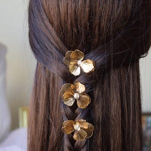May include: Close-up of a braid with three gold flower hair clips. Each clip has a small, clear gem in the center. The hair is dark brown and the clips are evenly spaced along the braid.
