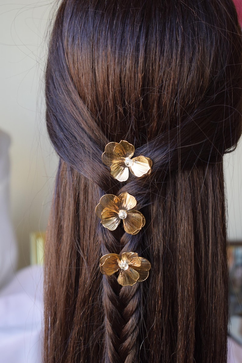 May include: Close-up of a braid with three gold flower hair clips. Each clip has a small, clear gem in the center. The hair is dark brown and the clips are evenly spaced along the braid.