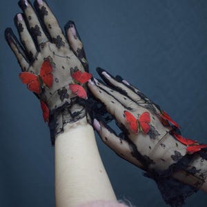 May include: Black lace gloves adorned with red butterfly embellishments. The gloves feature a floral pattern and a ruffled edge at the wrist. The butterflies add a whimsical touch to the elegant design.