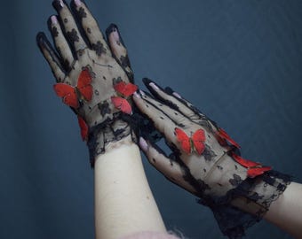 May include: Black lace gloves adorned with red butterfly embellishments. The gloves feature a floral pattern and a ruffled edge at the wrist. The butterflies add a whimsical touch to the elegant design.