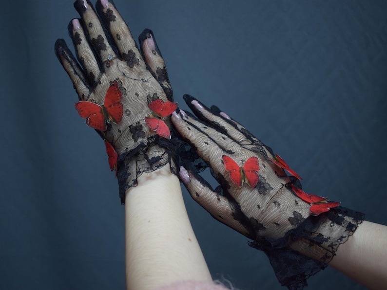 May include: Black lace gloves adorned with red butterfly embellishments. The gloves feature a floral pattern and a ruffled edge at the wrist. The butterflies add a whimsical touch to the elegant design.