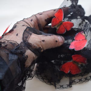 May include: Black lace gloves with a delicate polka dot pattern and scalloped edges. The gloves are adorned with three red butterfly appliqu&eacute;s, adding a pop of color to the elegant design. A red feather is also visible.