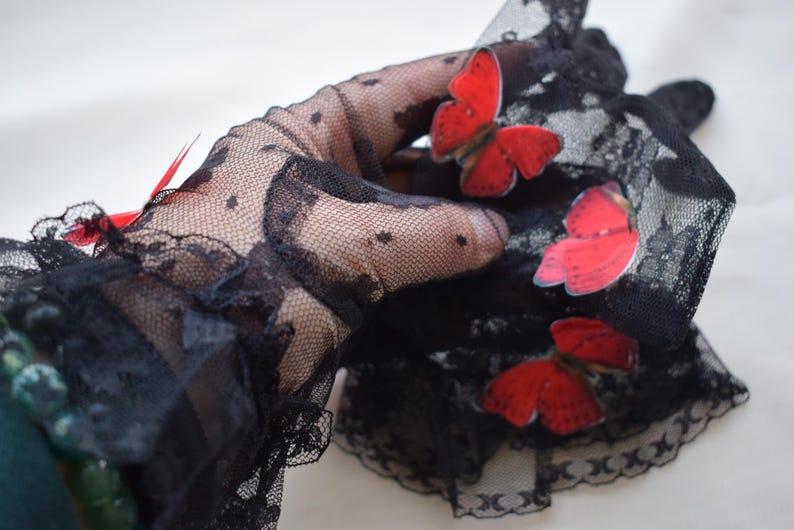 May include: Black lace gloves with a delicate polka dot pattern and scalloped edges. The gloves are adorned with three red butterfly appliqu&eacute;s, adding a pop of color to the elegant design. A red feather is also visible.