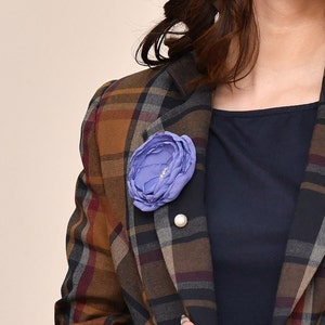 May include: A purple fabric flower brooch with a pearl accent pinned to a brown and black plaid blazer.