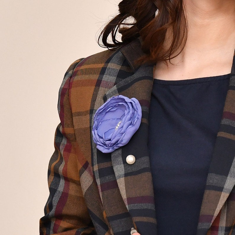 May include: A purple fabric flower brooch with a pearl accent pinned to a brown and black plaid blazer.