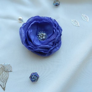 May include: A purple fabric flower brooch with pearl accents. The brooch is pinned to a white fabric background.
