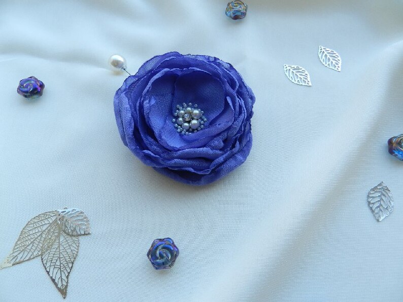 May include: A purple fabric flower brooch with pearl accents. The brooch is pinned to a white fabric background.