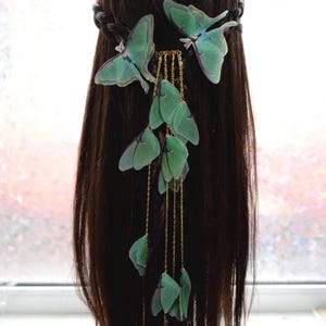 Könnte beinhalten: Ein Haarschmuck mit grünen Schmetterlingsakzenten. Die Schmetterlinge sind an goldenen Ketten befestigt, die das Haar hinunterfallen. Das Haar ist dunkelbraun und mit einem Zopf versehen.