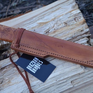 May include: A brown leather sheath with a knife inside. The knife has a wooden handle with a patterned design. The sheath has stitching around the edges. A black tag with the text "KAZAN KNIVES" is attached to the sheath.