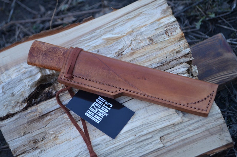 May include: A brown leather sheath with a knife inside. The knife has a wooden handle with a patterned design. The sheath has stitching around the edges. A black tag with the text "KAZAN KNIVES" is attached to the sheath.