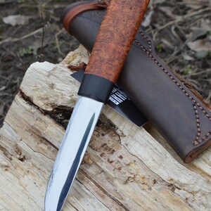 Puede incluir: Un cuchillo artesanal con una hoja de acero pulido y un mango de madera de ra&iacute;z. El cuchillo descansa sobre un trozo de madera, con una funda de cuero marr&oacute;n en el fondo. El mango mide unos 13 cm de largo.