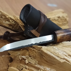 Könnte beinhalten: Ein kleines Messer mit schwarzem Griff, Holzgriff und Lederscheide. Das Messer liegt auf einem Stück Holz.
