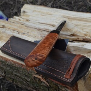 Puede incluir: Un cuchillo artesanal con mango de madera y funda de cuero. El cuchillo presenta una hoja oscura y un mango con un rico y ondulado veteado de madera. La funda de cuero marr&oacute;n tiene un borde cosido y una correa de cuero.