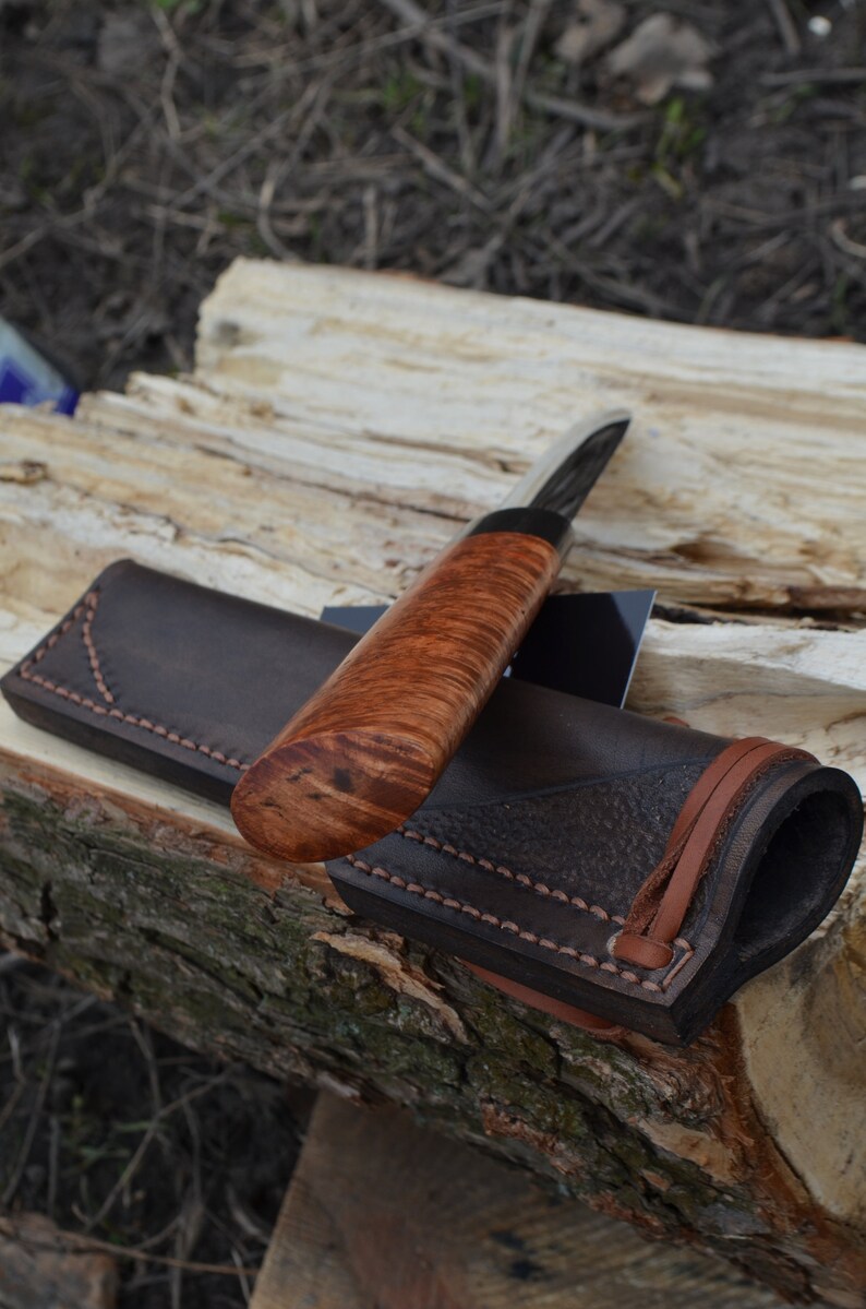 Puede incluir: Un cuchillo artesanal con mango de madera y funda de cuero. El cuchillo presenta una hoja oscura y un mango con un rico y ondulado veteado de madera. La funda de cuero marr&oacute;n tiene un borde cosido y una correa de cuero.