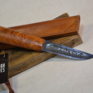 May include: A handmade knife with a wooden handle and a leather sheath. The blade is made of steel and has a hammered finish. The knife is resting on a wooden block with a tag that says "Kazhan Knives".