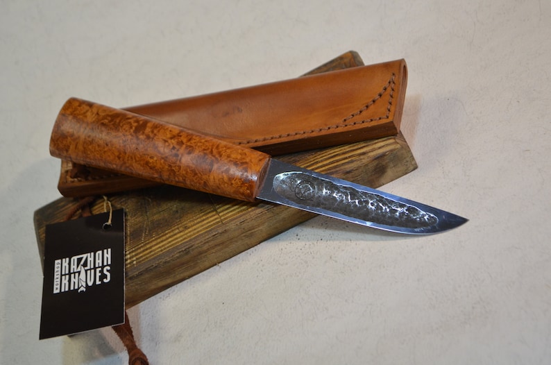 May include: A handmade knife with a wooden handle and a leather sheath. The blade is made of steel and has a hammered finish. The knife is resting on a wooden block with a tag that says "Kazhan Knives".