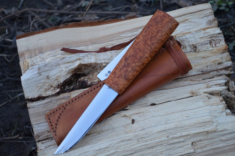 May include: A brown leather sheath with a brown wood handle knife with a silver blade. The knife is on a piece of wood.