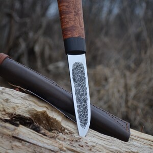 Puede incluir: Un cuchillo artesanal con una hoja texturizada y un mango de madera. El mango presenta una banda oscura cerca de la hoja. Se ve una funda de cuero marr&oacute;n. El cuchillo est&aacute; apoyado en un trozo de madera.