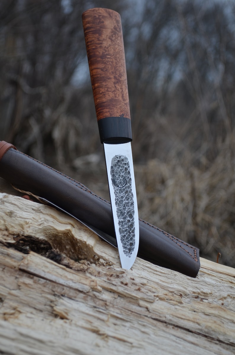 Puede incluir: Un cuchillo artesanal con una hoja texturizada y un mango de madera. El mango presenta una banda oscura cerca de la hoja. Se ve una funda de cuero marr&oacute;n. El cuchillo est&aacute; apoyado en un trozo de madera.