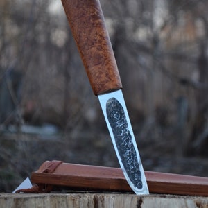 May include: A handcrafted knife with a wooden handle and a silver blade. The blade has a textured finish and is partially inserted into a brown leather sheath.