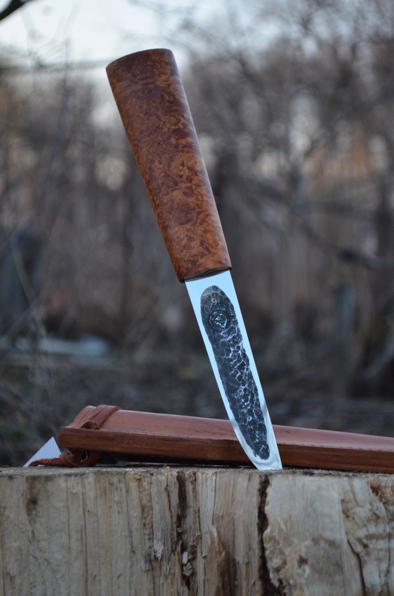 May include: A handcrafted knife with a wooden handle and a silver blade. The blade has a textured finish and is partially inserted into a brown leather sheath.