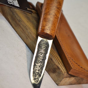 May include: A handmade knife with a brown wood handle and a leather sheath. The blade is made of steel and has a hammered finish. The knife is resting on a wooden block.