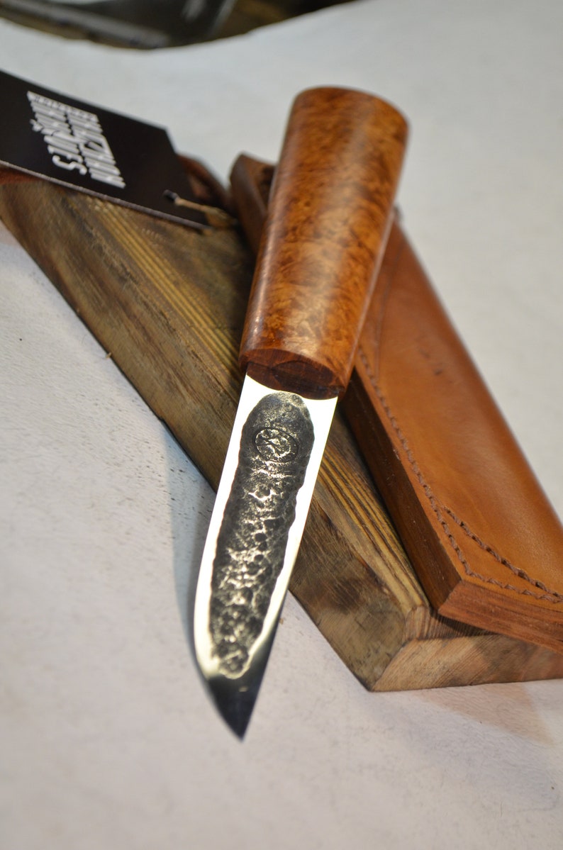 May include: A handmade knife with a brown wood handle and a leather sheath. The blade is made of steel and has a hammered finish. The knife is resting on a wooden block.