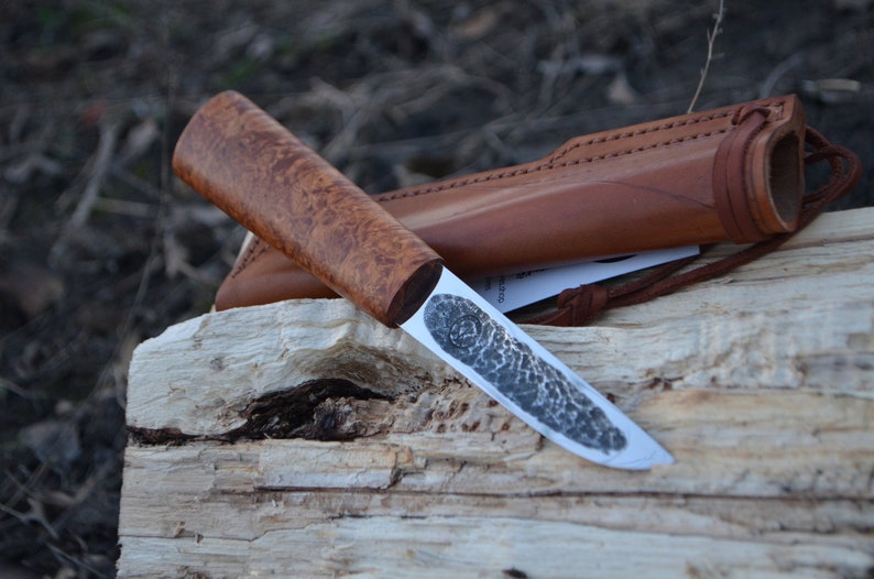 May include: A handmade knife with a wooden handle and a leather sheath. The blade is made of steel and has a hammered finish.