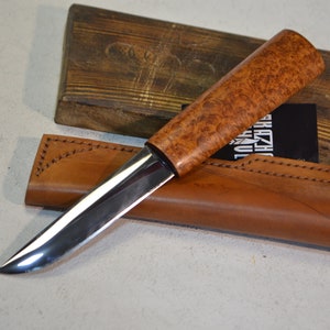 May include: A silver knife with a brown wooden handle and a leather sheath. The knife has a sharp point and a curved blade. The sheath has a brown leather strap with a knot at the end.