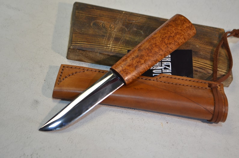May include: A silver knife with a brown wooden handle and a leather sheath. The knife has a sharp point and a curved blade. The sheath has a brown leather strap with a knot at the end.