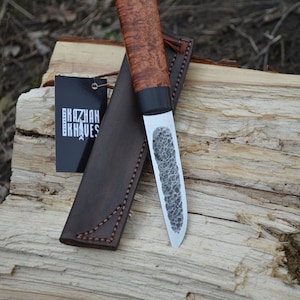 Puede incluir: Un cuchillo artesanal con una hoja texturizada y un mango de madera. El cuchillo va acompa&ntilde;ado de una funda de cuero marr&oacute;n y una etiqueta que dice "KAZHAN KNIVES". El cuchillo est&aacute; apoyado sobre un trozo de madera.