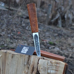 May include: A brown wooden handled knife with a silver blade. The blade has a textured finish. The knife is resting on a wooden stump with a leather sheath. The knife has a tag that says "Kizlar Knives".