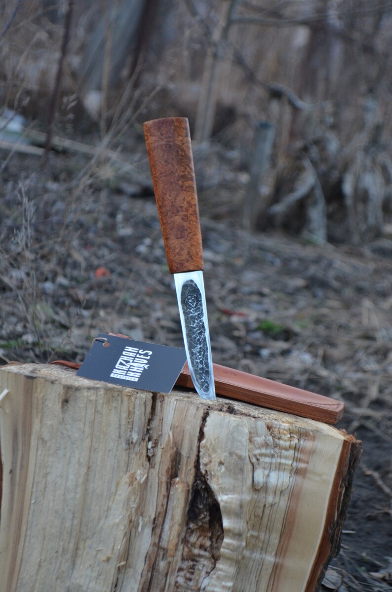 May include: A brown wooden handled knife with a silver blade. The blade has a textured finish. The knife is resting on a wooden stump with a leather sheath. The knife has a tag that says "Kizlar Knives".