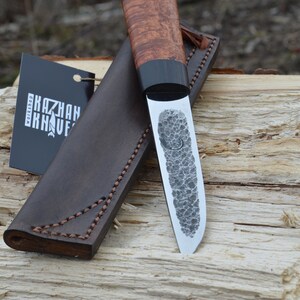 Puede incluir: Un cuchillo artesanal con una hoja estampada y un mango de madera. El cuchillo se muestra con una funda de cuero marr&oacute;n y una etiqueta que dice "KAZHAN KNIVES WORKSHOP". El mango tiene una banda oscura cerca de la hoja.
