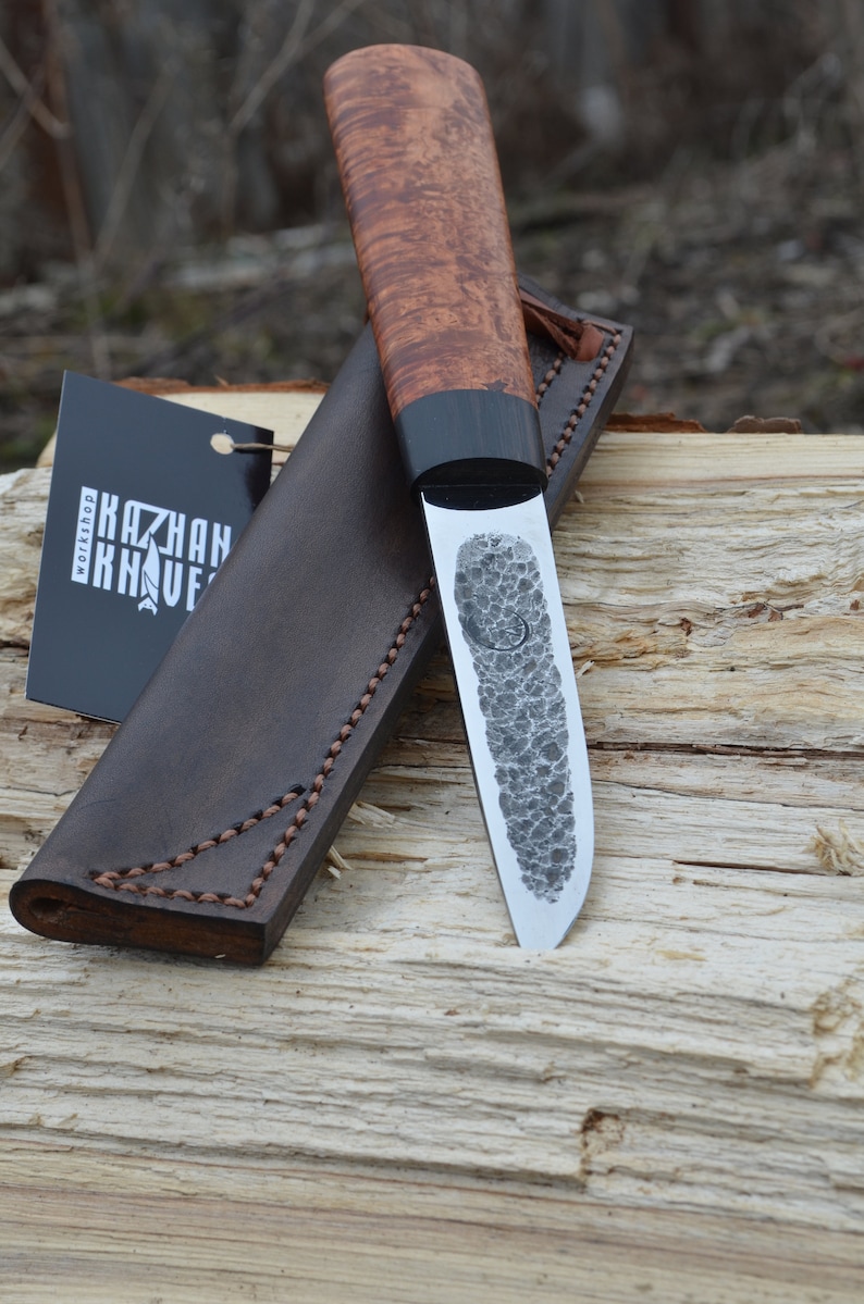 Puede incluir: Un cuchillo artesanal con una hoja estampada y un mango de madera. El cuchillo se muestra con una funda de cuero marr&oacute;n y una etiqueta que dice "KAZHAN KNIVES WORKSHOP". El mango tiene una banda oscura cerca de la hoja.