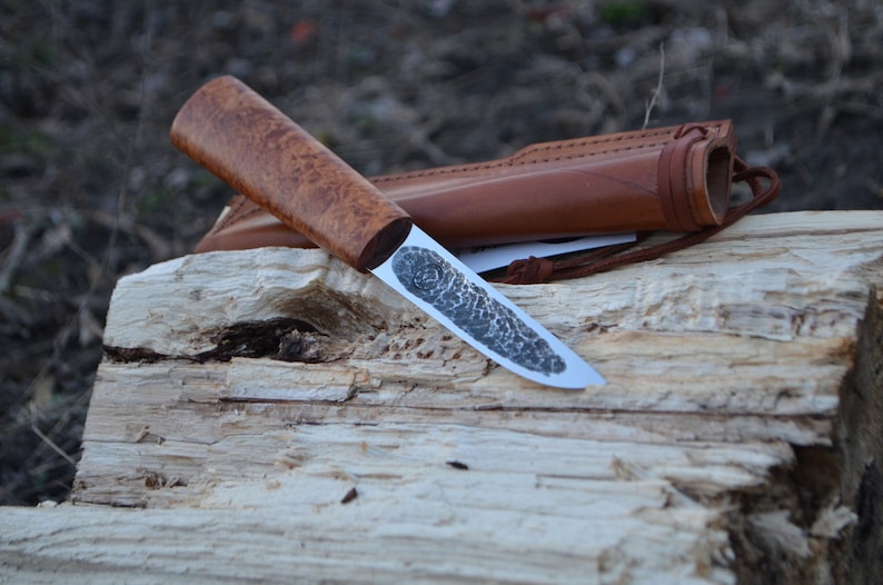 May include: A handmade knife with a wooden handle and a leather sheath. The blade is made of steel and has a hammered finish.