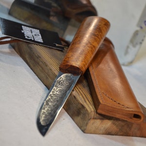 May include: A handmade knife with a hammered steel blade and a wooden handle. The knife is resting on a wooden surface with a leather sheath.