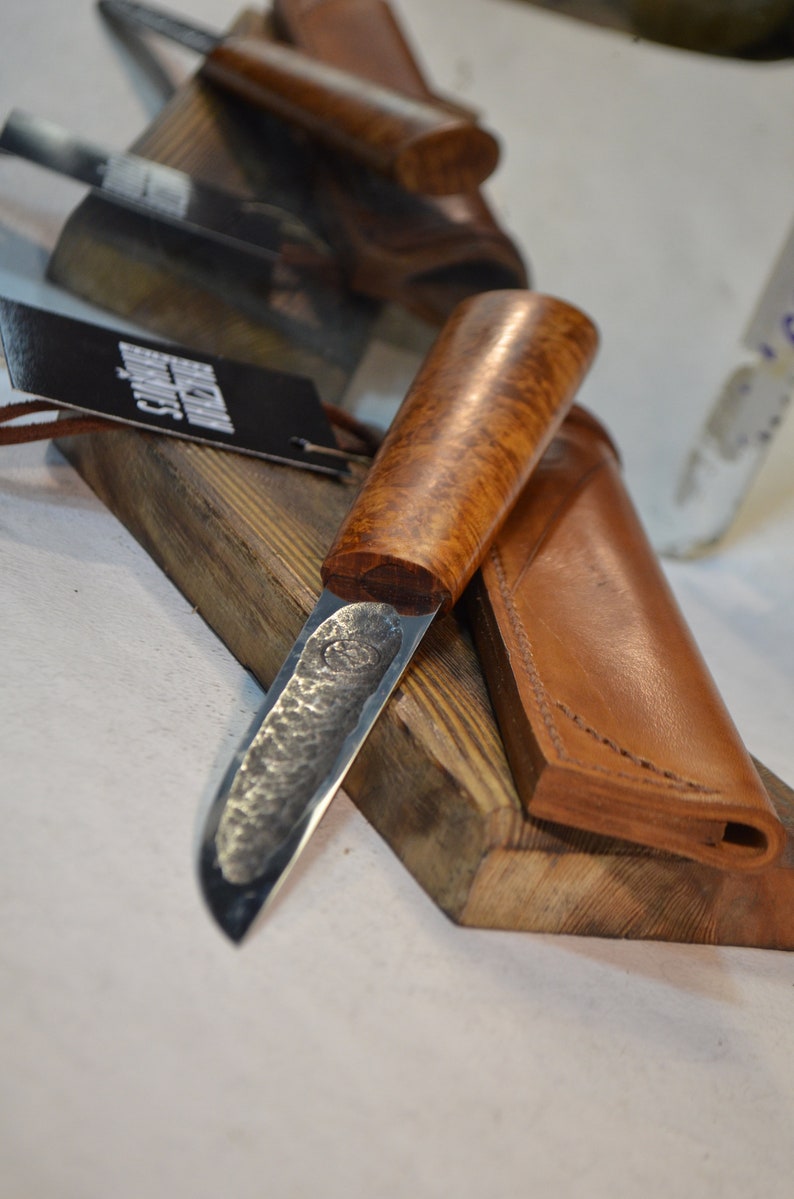 May include: A handmade knife with a hammered steel blade and a wooden handle. The knife is resting on a wooden surface with a leather sheath.