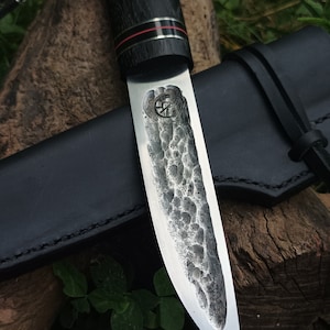 Könnte beinhalten: Ein Messer mit schwarzem Griff, silberner Klinge und strukturierter Oberfläche. Das Messer befindet sich in einer schwarzen Lederscheide mit einem braunen Lederriemen.